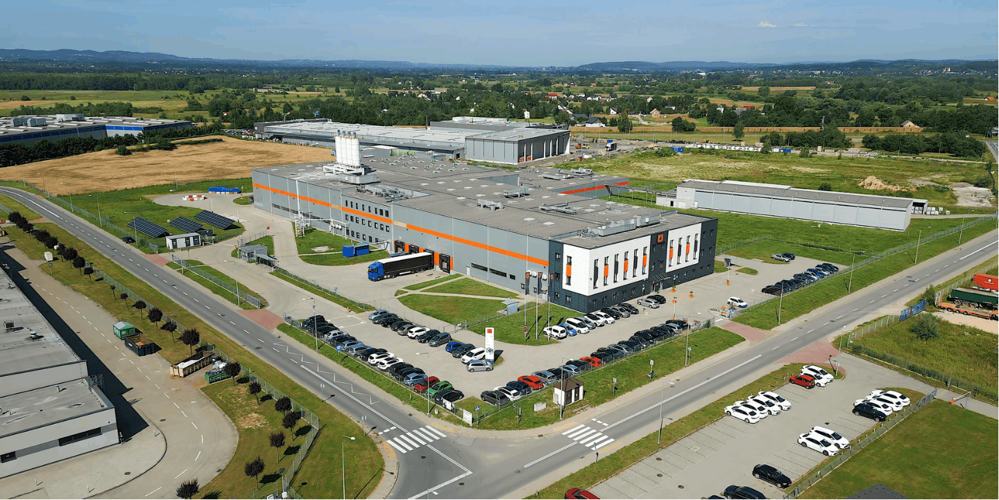 Widok z lotu ptaka na nowoczesny kompleks biurowo-produkcyjny firmy Dragon Poland w Skawinie. Rozległy, szaro-pomarańczowy budynek z widocznym parkingiem, strefą logistyczną z dokami załadunkowymi oraz instalacją fotowoltaiczną, otoczony terenami zielonym